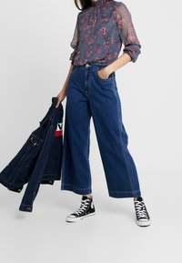 Květnatá šedá blůza s průhledným materiálem a vyboulenými rukávy, doplněná o široké modré džíny a černé vysoké tenisky. Džínová bunda držena v ruce.