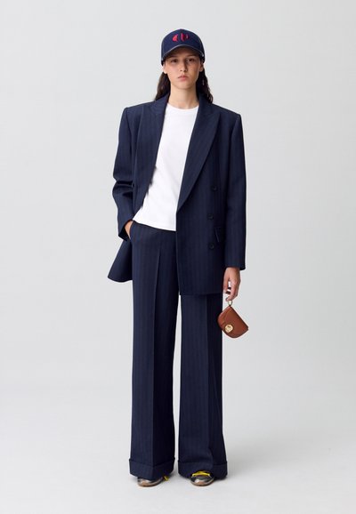 Traje a rayas marinas que incluye un blazer de doble botonadura, pantalones de pierna ancha, camiseta blanca, gorra marina con logo y un bolso mini marrón.