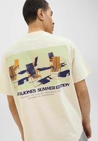 Short-sleeve cream t-shirt featuring a graphic of striped deck chairs in blue and orange on a green background with text.