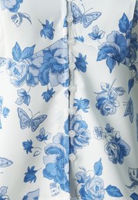 Hvit bluse i stoff med et blått blomstermønster, sommerfugler og runde hvite knapper foran. Glatt tekstur og tettsittende design.