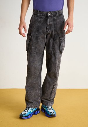 Person trägt gewaschene schwarze Cargo-Hose und blau-schwarze-lila Sportschuhe und steht auf einem gelben Boden.