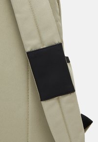 Correa de mochila beige con acento negro. El material parece ser una tela duradera con una superficie texturizada. Presenta bordes cosidos y una pestaña negra.