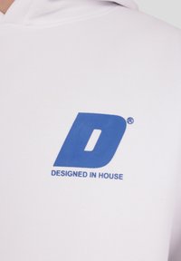 Biela mikina s kapucňou s veľkým modrým logom obsahujúcim písmeno „D“ a textom „DESIGNED IN HOUSE“ potlačeným na hrudi. Hladká textúra.