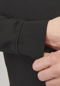 Ingrandimento di una mano che aggiusta la manica di una felpa nera con polsino a costine su un braccio di tonalità della pelle chiara.