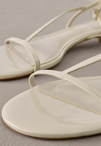 Gros plan de sandales crème avec fines brides et design à bout carré sur une surface neutre.