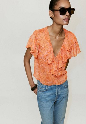 Femme portant de grandes lunettes de soleil foncées, un chemisier orange à motifs avec des volants, un jean bleu et un bracelet sombre à son poignet.