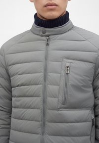 Homme portant une veste matelassée gris clair avec un col montant et une poche poitrine verticale zippée sur un pull à col roulé côtelé foncé.