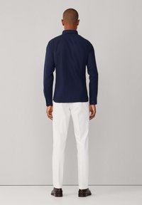 Camicia a maniche lunghe color blu navy realizzata in tessuto liscio, con collo button-down e polsini con più bottoni, abbinata a pantaloni bianchi su misura.