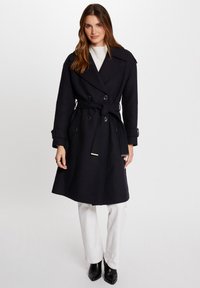 Marineblauwe dubbelgebreide jas met een riem, met zijzakken en knoopaccenten. Model draagt witte broeken en zwarte hoge laarzen.
