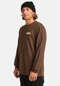 Camisa marrón de manga larga en tela suave y transpirable con cuello redondo y puños acanalados; presenta un parche con el logo en el pecho.