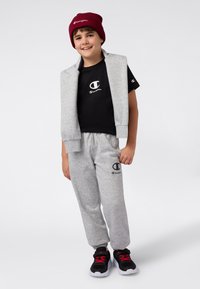 Pantalones de chándal grises y una sudadera sin mangas gris sobre una camiseta negra, combinados con zapatos negros y rojos y un gorro burdeos.