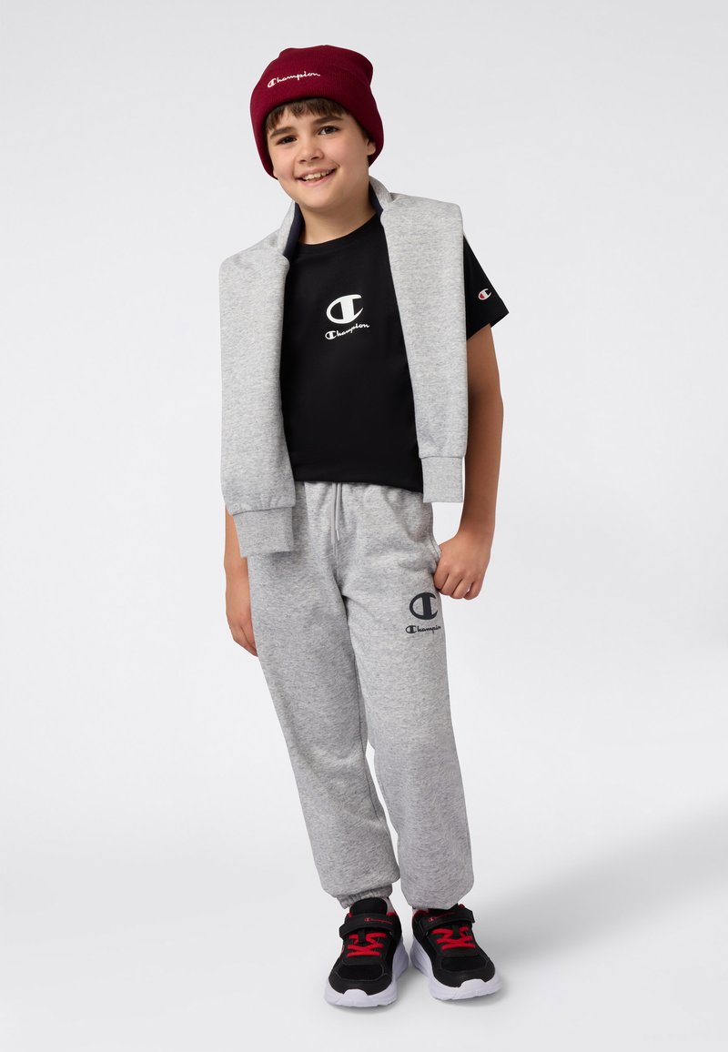 Pantalones de chándal grises y una sudadera sin mangas gris sobre una camiseta negra, combinados con zapatos negros y rojos y un gorro burdeos.