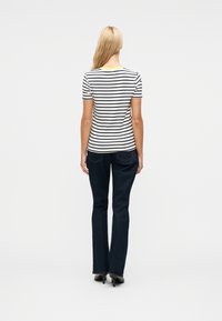 Γυναίκα με μακριά ξανθά μαλλιά, ντυμένη με λευκή και μαύρη ριγέ κοντομάνικη μπλούζα, σκούρο μπλε τζιν και μαύρα παπούτσια με τακούνι, στεκόμενη με την πλάτη προς τα εμπρός.