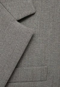 Tissu gris texturé avec un motif de tissage visible et un détail de poche. Présente des bords de col nets. Aucun ornement ni quincaillerie présents.