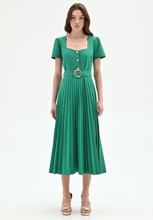 Donna che indossa un vestito midi verde a maniche corte, abbottonato, con gonna plissettata e cintura coordinata con fibbia decorativa, abbinato a tacchi dorati.