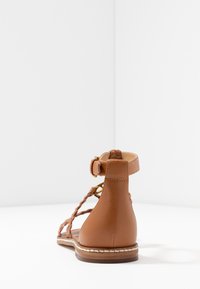 Sandale en cuir marron avec des brides entrelacées, fermeture à la cheville et un petit talon. Dispose d'une texture lisse et d'un détail de couture blanche contrastant.