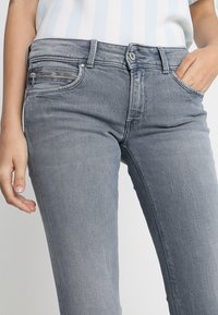Los pantalones vaqueros de mezclilla gris tienen un corte ajustado, bolsillos frontales con acentos de cremallera y una cinturilla clásica. La tela tiene una textura suave y lisa.