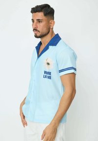 Chemise à manches courtes bleu clair avec un col bleu marine, ornée d'un motif de fleur blanche et du texte "VIVRE LA VIE" en bleu.