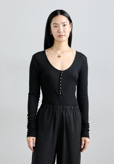 Sort langærmet bodysuit med dyb V-hals og knaplukning. Blødt materiale, tætsiddende design og ribbet tekstur, parret med høj talje sorte bukser.