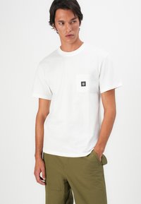 Young man wearing a plain white T-shirt with a black square logo on the chest pocket and olive green pants, standing with one hand in pocket.