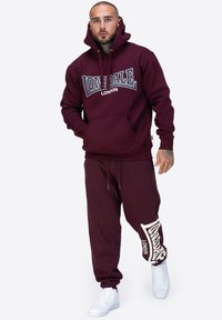 Burgundowa bluza z kapturem z przednią kieszenią i logo, zestawiona z pasującymi dresami z białym logo na nogawce, uzupełniona białymi sneakersami.