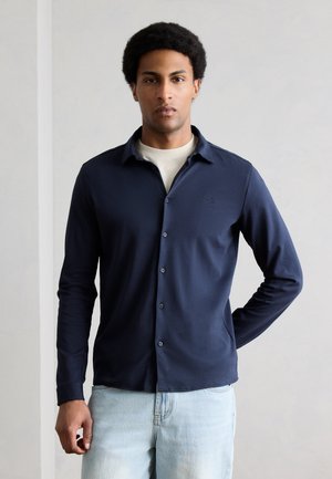 Άνδρας που στέκεται μπροστά σε απλό τοίχο, φοράει μπλε πουκάμισο με κουμπιά πάνω από κρεμ T-shirt και ανοιχτό μπλε τζιν, με το ένα χέρι πίσω από την πλάτη.