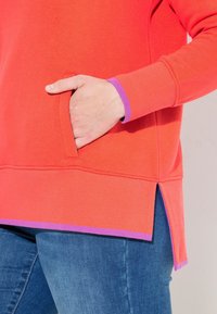 Sudadera de color coral con un bolsillo lateral, acentos morados en los puños y el dobladillo, textura acanalada y laterales abiertos. Combinada con jeans azules.