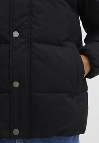 Veste rembourrée noire avec boutons pression, présentant une texture matelassée et des poches latérales. Comprend un patch de logo de la marque au bas.