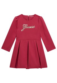 Robe en coton rouge à manches longues avec une jupe plissée, présentant un logo "Guess" pailleté sur la poitrine et une fermeture éclair dans le dos.
