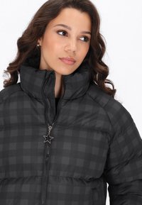 Černá a šedá kostkovaná péřová bunda s vysokým límcem a zipovým zapínáním, na kterém visí hvězdný přívěsek. Hladká tkanina s prošívanou strukturou.