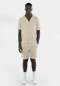 Lys beige kortermet skjorte og shorts-sett laget av teksturert stoff. Har knapper og en avslappet passform. Kombinert med hvite joggesko og sokker.