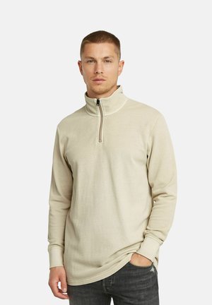 Beiger Zip-Pullover mit langen Ärmeln und einem strukturierten Stoff. Der Kragen ist hoch und die Passform ist lässig, kombiniert mit dunklen Jeans.