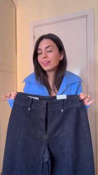Fiatal nő kék pulóverben egy pár fekete farmert tart maga előtt, bézs szobabelső háttér előtt.