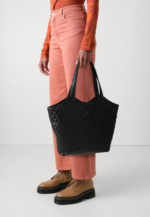 Personne portant des bottes marron clair et un pantalon rose saumon tenant un grand sac cabas noir matelassé avec poignées.