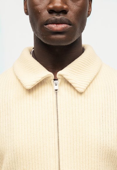 Κοντινό πλάνο ενός ατόμου που φοράει πλεκτό μπουφάν εκρού με φερμουάρ και φαρδύ γιακά, πάνω σε απλό φόντο.