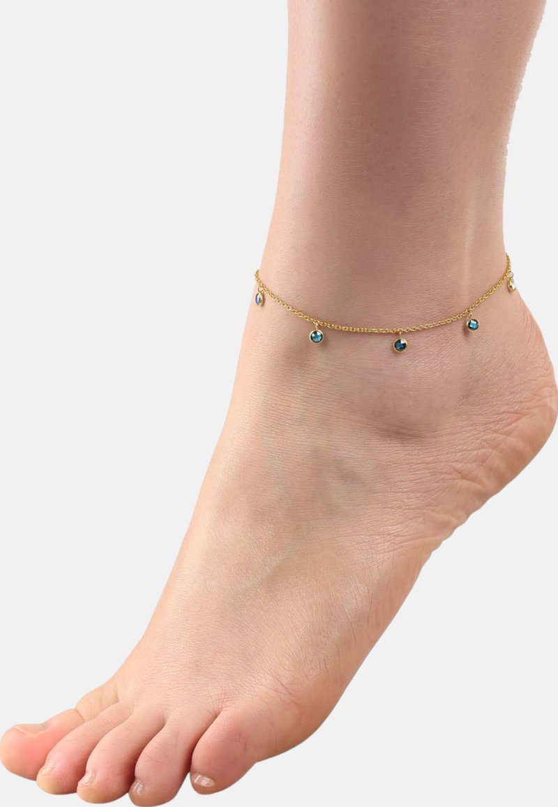 Tobillera con cadena de oro que presenta pequeñas cuentas azules redondas. La cadena tiene un diseño delicado, envolviendo el tobillo. Se ve un pie de tono de piel.