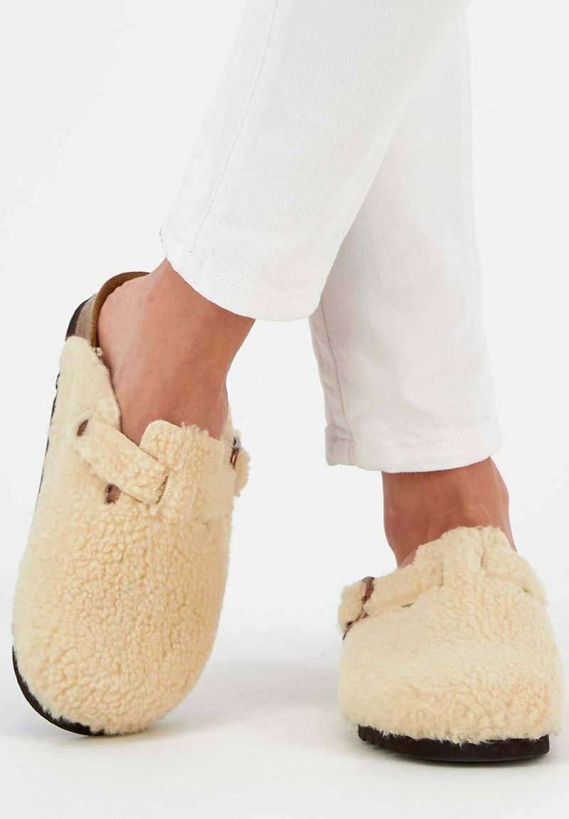 Fuzzy cream slippers with a soft, textured finish, featuring a wide strap and dark sole. Worn with white cropped pants.