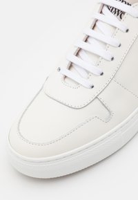 Bílé kožené tenisky s hladkým povrchem, vyznačující se polstrovaným jazykem, bílými tkaničkami a texturovanou gumovou podrážkou. Žádná viditelná loga.