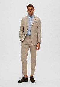 Lichtbeige pak met een gestructureerde afwerking, gecombineerd met een lichtblauw overhemd met knopen. De broek heeft een slim-fit, aangevuld met donkere schoenen.