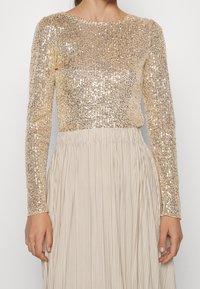 Gouden pailletten top met lange mouwen en een ronde halslijn, gedragen met een geplisseerde beige rok, wat een gelaagd en elegant uiterlijk creëert.