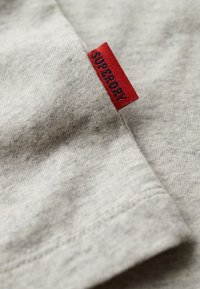 Tessuto grigio chiaro con superficie testurizzata, caratterizzato da un'etichetta rossa con "SUPERDRY" in blu scuro, che indica il brand.