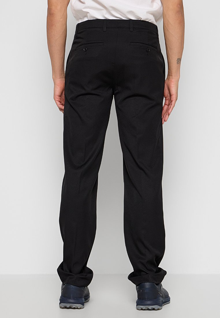 Homme portant un pantalon noir à coupe droite avec des poches arrière à boutons et des chaussures bleu marine, se tenant contre un fond blanc uni.