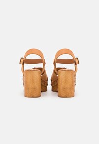 Sandales en cuir tan avec des semelles compensées épaisses en bois et des brides autour de la cheville, vues de l'arrière sur un fond blanc.