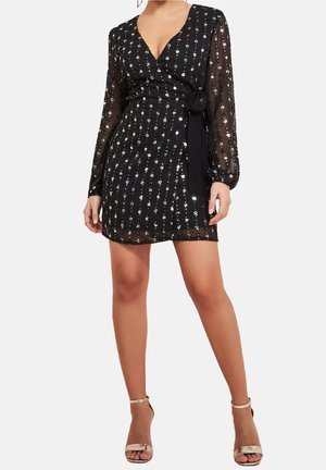 Czarna mini sukienka kopertowa z przezroczystymi długimi rękawami i pionowymi srebrnymi cekinami, w zestawieniu ze złotymi szpilkami z paskiem wokół kostki.