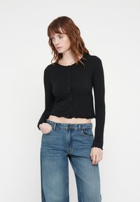 Svart kortermet cardigan med langermede, knappelukking, med bølgete kanter og en myk tekstur, kombinert med høyt midje blå jeans.