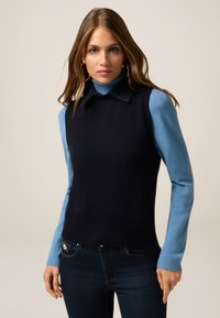 Marineblauw mouwloos gebreid topje met ribbelrand en kraag, gelaagd over een lichtblauw gebreid turtleneck met lange mouwen. Donkerdenim jeans.