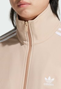 Beige zip-up jacket with high collar, featuring white three-stripe accents and an embroidered logo on the chest, made from smooth material.