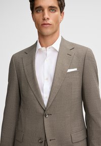 Męska dopasowana brązowa marynarka w kratę z klapami z wycięciem, dwoma guzikami i białą poszetką, noszona na białej koszuli wizytowej.