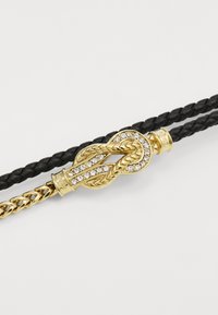 Breda läderarmband i guld och svart med ett dekorativt guldspänne med klara stenar och repliknande detaljer.