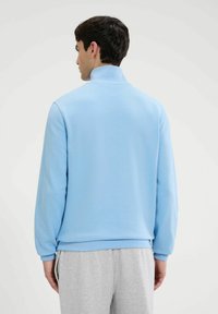 Sudadera de forro polar azul claro con cuello alto, puños acanalados y dobladillo. Presenta una textura suave y un corte holgado. Combinada con pantalones de chándal grises.